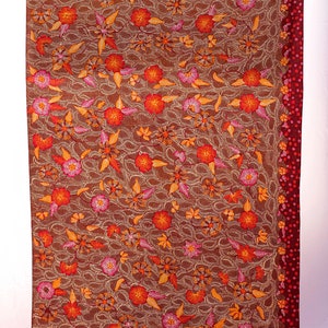 May include: Brown textile with an intricate floral pattern in shades of orange, pink, and gold. The design features detailed embroidery of flowers and leaves. A deep red border with small dots runs along one edge.