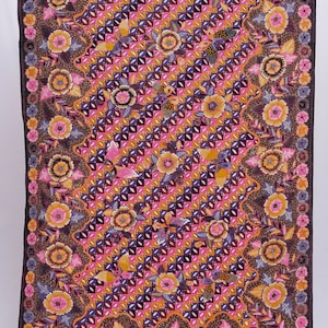 Könnte beinhalten: Rechteckiger Stoff mit einem lebendigen Blumenmuster. Das Design zeigt ein sich wiederholendes geometrisches Muster in Rosa, Orange und Lila, mit einem Rand aus Blumen in Rosa-, Gelb- und Brauntönen. Der Stoff ist wahrscheinlich ein Schal oder ein dekoratives Stück.