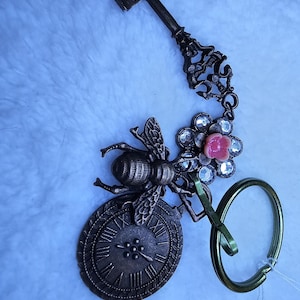 Puede incluir: Un llavero de bronce con una abeja, una esfera de reloj y una flor rosa con pedrería. El llavero está unido a un anillo de metal verde.