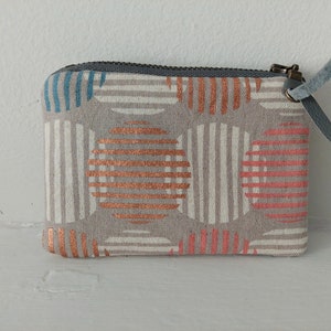 May include: A small grey zippered pouch with a white background and a pattern of orange and pink stripes in circles. The zipper pull is a brown metal with a grey leather tab.