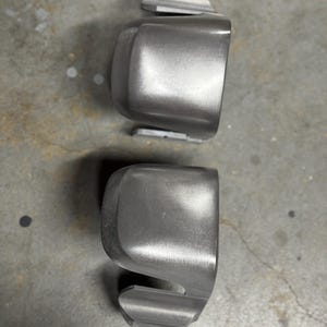 May include: Two grey, rounded automotive parts are shown against a grey surface. Each part has a curved body with a rectangular extension on top and a smaller, angled piece at the bottom. The parts appear to be made of metal.