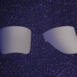 May include: Two smooth, light gray, curved plastic visor components. One piece is shown from a front-facing perspective, while the second piece is positioned at an angle to display its side profile and curved shape. Both items have a matte finish.