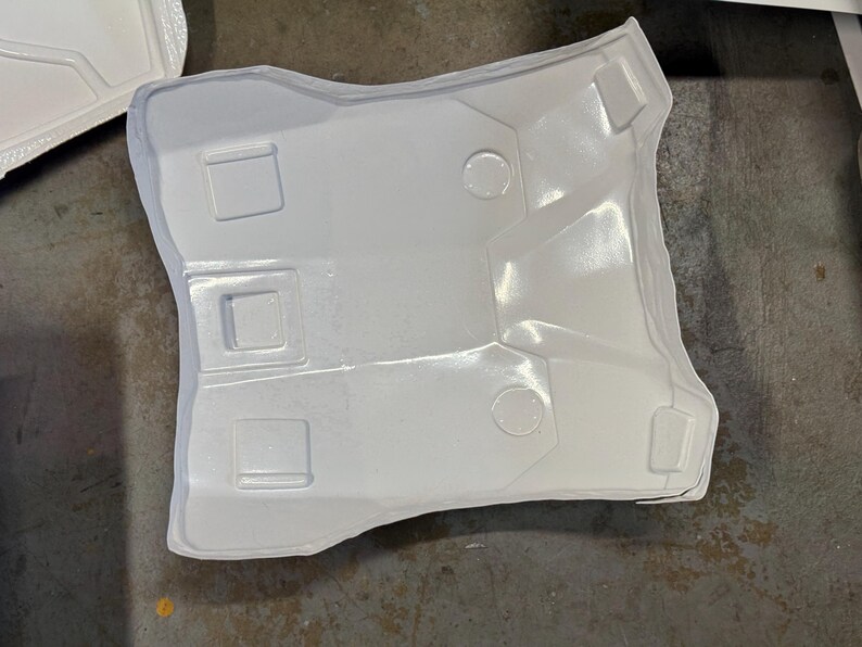 Puede incluir: Un componente de pl&aacute;stico moldeado blanco con forma curva y detalles geom&eacute;tricos. La pieza presenta tres hendiduras rectangulares y dos circulares, con una superficie texturizada. El art&iacute;culo parece ser parte de una estructura m&aacute;s grande.