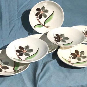 Pode incluir: Conjunto de oito tigelas de cerâmica branca com um design floral. As tigelas apresentam uma flor estilizada com folhas verdes e pétalas marrons e cinzas.