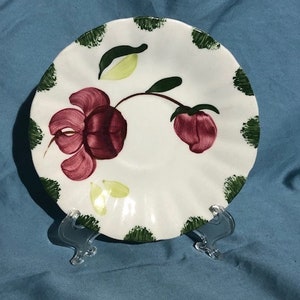 Puede incluir: Un platillo de cerámica blanco con un diseño floral. El platillo presenta una flor y un capullo granate con hojas y detalles verdes. El borde está decorado con pinceladas verdes. El platillo se exhibe sobre un soporte de plástico transparente.