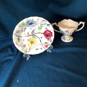 Może przedstawiać: Biały ceramiczny spodek z kwiatowym wzorem w odcieniach niebieskiego, żółtego, czerwonego i zielonego. Na zdjęciu znajduje się również pasująca biała ceramiczna filiżanka z dwoma uchwytami.