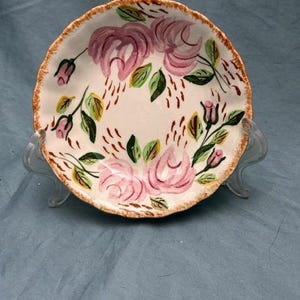 May include: A small, round ceramic plate with a white base, decorated with hand-painted pink roses and green leaves. The rim has a brown, textured edge. The plate is displayed on a clear stand, showcasing its decorative floral design.