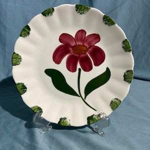 Puede incluir: Plato de cerámica blanca con borde festoneado, con una flor roja pintada a mano con centro amarillo y hojas verdes. El borde tiene lunares verdes. El plato se exhibe sobre un soporte de plástico transparente.