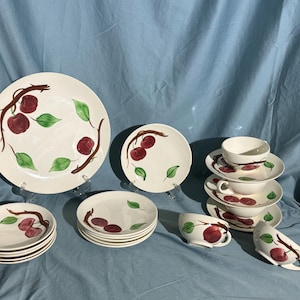 May include: A vintage set of white ceramic dinnerware featuring a red and green cherry design. The set includes a large platter, salad plates, teacups, and saucers.