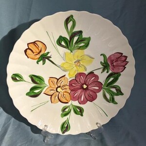Puede incluir: Un plato blanco con borde festoneado y diseño floral pintado a mano. Las flores son en tonos amarillo, naranja y burdeos, con hojas verdes. El plato se exhibe sobre un soporte transparente, mostrando una estética vintage.