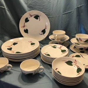 May include: A set of vintage white ceramic dinnerware with a green and red floral design. The set includes plates, bowls, and cups.