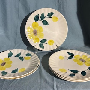 May include: A collection of white ceramic plates featuring a yellow floral pattern. The plates have a scalloped edge and are adorned with yellow flowers, green leaves, and yellow buds. The plates are presented on a blue surface.