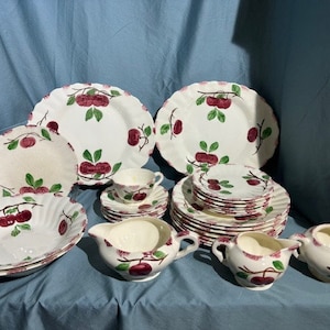 May include: A vintage set of white ceramic dinnerware featuring a cherry design. The set includes plates, bowls, and a creamer and sugar bowl.