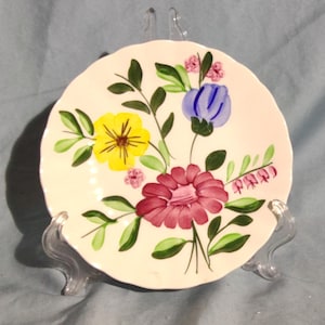 Puede incluir: Un plato redondo de cerámica de color crema con borde festoneado, que presenta un diseño floral pintado a mano. El patrón incluye una flor amarilla grande, una flor rosa oscuro prominente, una flor azul y pequeñas flores rosas con hojas verdes.