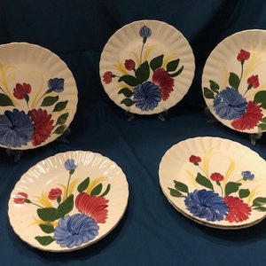 May include: Set of five white ceramic dinner plates with a floral design. The plates feature a blue and red flower with green leaves and yellow stems. The plates have a scalloped edge.