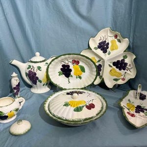 May include: A vintage set of ceramic dishes featuring a fruit motif. The set includes a teapot, creamer, covered dish, and three serving dishes. The dishes are decorated with grapes, pears, and cherries in shades of purple, green, yellow, and red.
