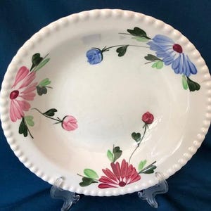 May include: A white ceramic bowl with a scalloped edge and a floral design. The bowl features pink, blue, and green flowers with green leaves.