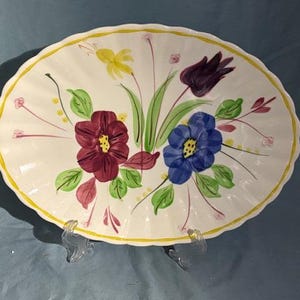 May include: Oval ceramic serving dish with a scalloped edge and floral design. The flowers are painted in shades of burgundy, blue, and yellow, with green leaves and stems. The rim is trimmed with a yellow border.
