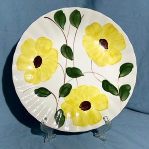 May include: A white ceramic plate with a yellow floral design. The plate features three yellow flowers with brown centres and green leaves.