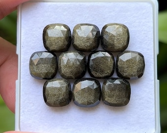 Pode incluir: Nove pedras preciosas de obsidiana dourada, lapidadas em forma quadrada e facetadas, são exibidas numa bandeja branca. As pedras têm um brilho metálico refletor e estão dispostas em três filas de três. O fundo está desfocado.