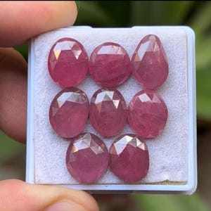 May include: A collection of eight faceted, oval and pear-shaped, deep pink gemstones. The gems are displayed in a white plastic tray, held by a hand. The gems have a polished, reflective surface, and are likely rubies.