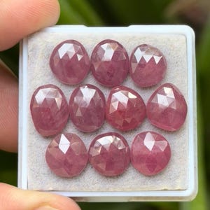 May include: A collection of ten faceted, oval-shaped, pink gemstones displayed in a white tray. The gemstones have a rose-like hue and are arranged in three rows. The image is well-lit, showcasing the gems' clarity and cut.