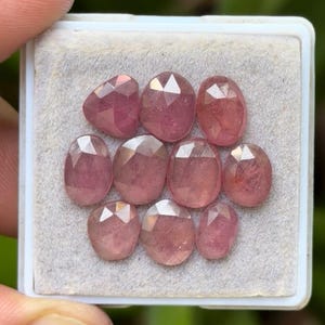 May include: A collection of ten faceted, pink gemstones displayed in a white plastic tray. The gemstones vary in shape, including oval and rounded forms. The gems have a translucent quality, with visible internal structures.