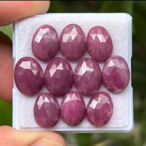 Puede incluir: Una colección de nueve piedras preciosas de rubí ovaladas y facetadas. Los rubíes son de un color rojo-rosado intenso y se muestran en una bandeja blanca. Las facetas crean un efecto brillante, resaltando la belleza natural de las piedras.