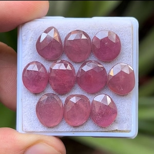 May include: A collection of ten faceted, oval-shaped gemstones in a light pink hue, arranged in a white plastic tray. The gemstones have multiple facets that catch the light, creating a sparkling effect. The gems are likely rubies or pink sapphires.
