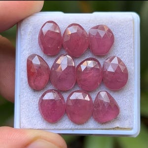 May include: A collection of ten faceted, oval-shaped gemstones in varying sizes, displayed in a white tray. The gems are a deep pinkish-red color, with multiple facets catching the light. The gems are held by a hand.