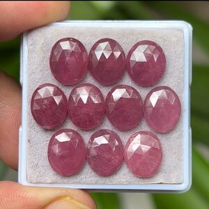 May include: Ten oval-shaped, faceted gemstones in a light pink hue are arranged in a rectangular, light grey container. The gemstones have a polished, reflective surface, and are displayed against a blurred green background.