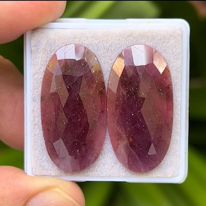 Könnte beinhalten: Zwei ovale, facettierte Edelsteine in tiefem Rot-Violett. Die Steine werden nebeneinander in einem weißen Kunststoffbehälter präsentiert. Die Facetten fangen das Licht ein und erzeugen einen funkelnden Effekt. Die Steine sind wahrscheinlich für die Schmuckherstellung bestimmt.