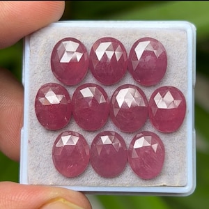 May include: Ten oval-cut gemstones in a light pink colour, displayed in a rectangular plastic container. The faceted stones have a polished surface, reflecting light. They are set against a light grey background.