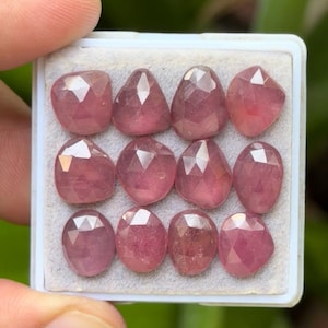 May include: A collection of twelve faceted, pink gemstones displayed in a white tray. The gemstones have various shapes, including oval, teardrop, and square, and are held by a person's fingers.
