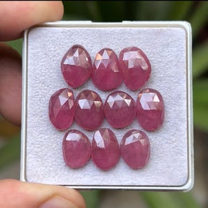 May include: A collection of nine faceted, oval-shaped, pink gemstones arranged in a white tray. The gems have a polished, reflective surface, and are a deep, rich shade of pink. The tray is held in a person's hand.