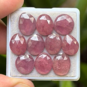 May include: A collection of ten faceted, oval-shaped pink gemstones displayed in a white tray. The gemstones have a translucent appearance and a polished finish, with multiple facets catching the light. The color is a soft, rosy pink.