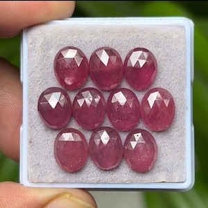 May include: A collection of nine oval-cut, faceted rubies displayed in a white tray. The gemstones are a deep, rich red color with multiple facets that catch the light. The rubies are set against a light gray background.