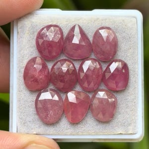 May include: A collection of ten faceted, pink gemstones displayed in a white tray. The gemstones have various shapes, including oval, teardrop, and round, and are set against a light gray background. The gemstones are held by a person's fingers.