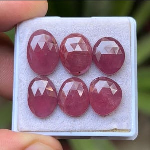 May include: Six faceted, oval-shaped, ruby gemstones displayed in a white tray. The rubies have a deep red color and a brilliant, multi-faceted cut that reflects light. The gemstones are arranged in two rows of three.