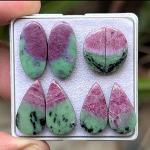 May include: A set of six green and pink gemstone cabochons in various shapes, including oval and teardrop. The stones are arranged in a white display box.
