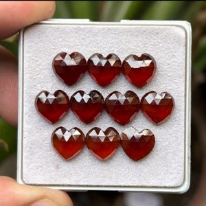 May include: Nine heart-shaped gemstones with faceted surfaces are arranged in a white tray. The gems are a deep red color, with a sparkling appearance. The tray is held by a person's hand, with a blurred background.