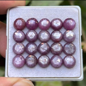 May include: A collection of sixteen faceted, round gemstones in a white tray. The gems are a deep pinkish-purple color, with multiple facets catching the light. The gems are arranged in four rows of four.