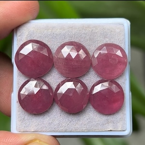Puede incluir: Seis gemas rosas redondas y facetadas se exhiben en una caja de plástico azul claro. Las gemas tienen una superficie pulida y reflectante y un color rosa intenso. Las gemas están dispuestas en dos filas de tres.