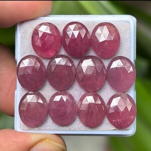 Puede incluir: Una colección de diez gemas ovaladas y facetadas en un tono rosa rojizo claro. Las gemas están dispuestas en una bandeja blanca rectangular, cada una mostrando un corte geométrico complejo que refleja la luz. El fondo es un verde suave y borroso.