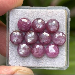 May include: A collection of ten faceted, round, pinkish-red gemstones. The gems are displayed in a clear plastic case with a gray interior. The gems have a brilliant cut, reflecting light and showcasing their internal structure.