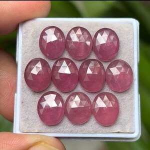 Puede incluir: Una colección de nueve piedras preciosas facetadas de forma ovalada en un tono rosa claro. Las gemas están dispuestas en una bandeja blanca, mostrando sus superficies pulidas y cortes intrincados. Las piedras preciosas son sostenidas por una mano.