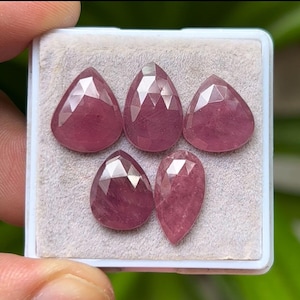 Op de afbeelding: Vijf gefacetteerde, druppelvormige edelstenen in verschillende maten. De edelstenen hebben een dieproze-rode kleur en worden gepresenteerd in een kleine, witgrijze container. De edelstenen hebben een gepolijst, reflecterend oppervlak.
