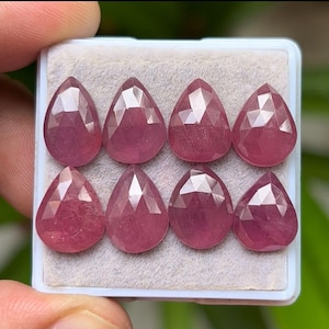 Op de afbeelding: Acht gefacetteerde, druppelvormige edelstenen in een lichtroze tint zijn gerangschikt in een wit dienblad. De edelstenen hebben een gepolijst, reflecterend oppervlak dat hun interne structuur laat zien. De lade wordt door een hand vastgehouden, met een vage groene achtergrond.