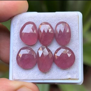 May include: Six faceted, oval-shaped gemstones in a white tray. The gems are a deep pink color, with multiple facets that catch the light. The gems are displayed on a white surface.