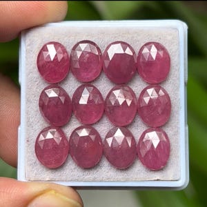 May include: A collection of twelve oval-cut, faceted gemstones in a light-colored tray. The gems are a deep pinkish-red color, with multiple facets catching the light. The gems are arranged in three rows of four.